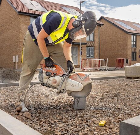 A groundsworker in a safety vest and helmet uses a power saw on a concrete block at a residential site. New build homes are visible in the background.