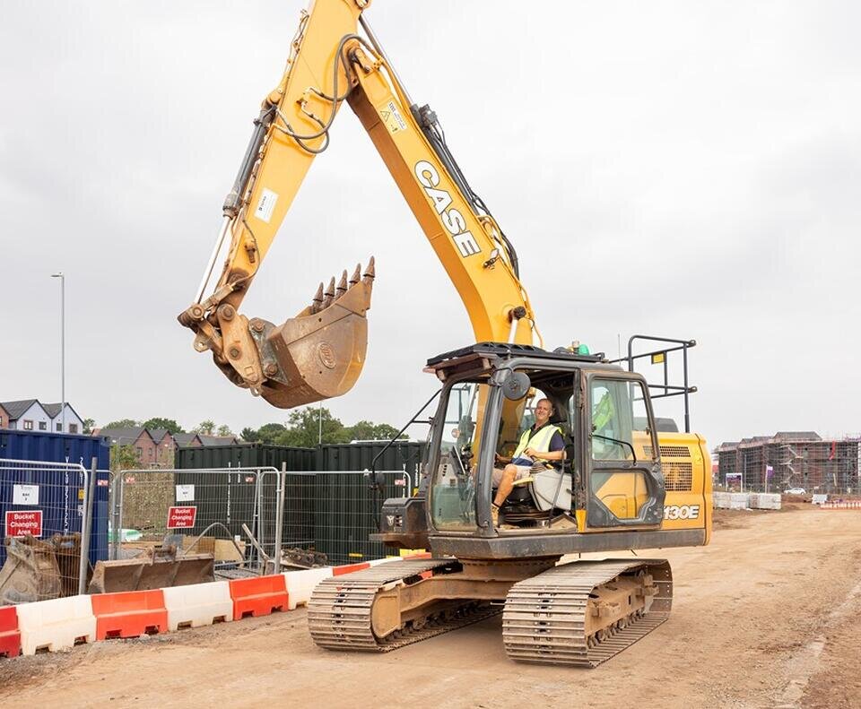 A yellow excavator operates on a construction site, surrounded by metal fencing and barriers. Overcast sky looms, conveying a serious work environment.
