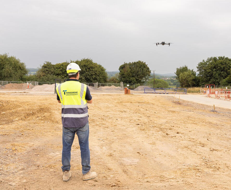 A groundsworker in a high vis vest and hard hat is piloting a drone over an expansive area of earth