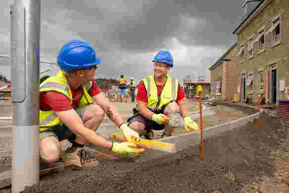 Two groundsworkers are kneeling down, laying new kerbs on a footpath which is yet to be complete. They have a spirit level and string to ensure it's level