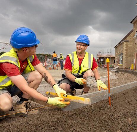 Two groundsworkers are kneeling down, laying new kerbs on a footpath which is yet to be complete. They have a spirit level and string to ensure it's level