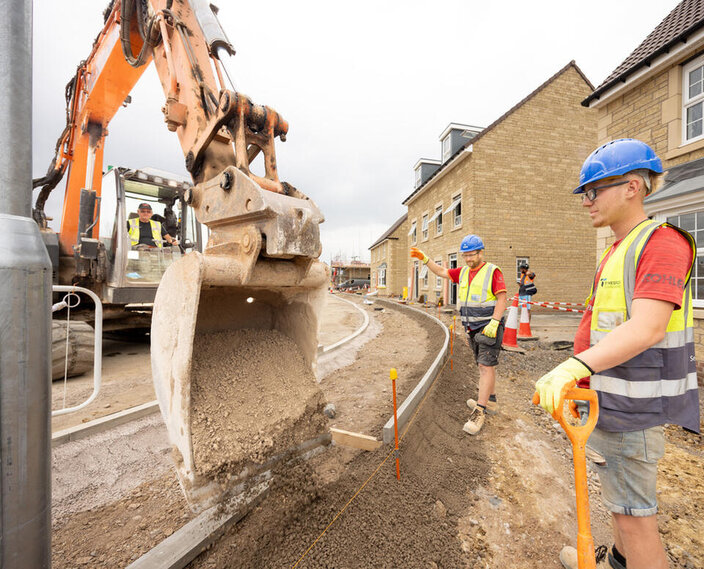 Two groundworkers are stood on the right directing a large digger which is pouring concrete onto a new footpath which is being laid.