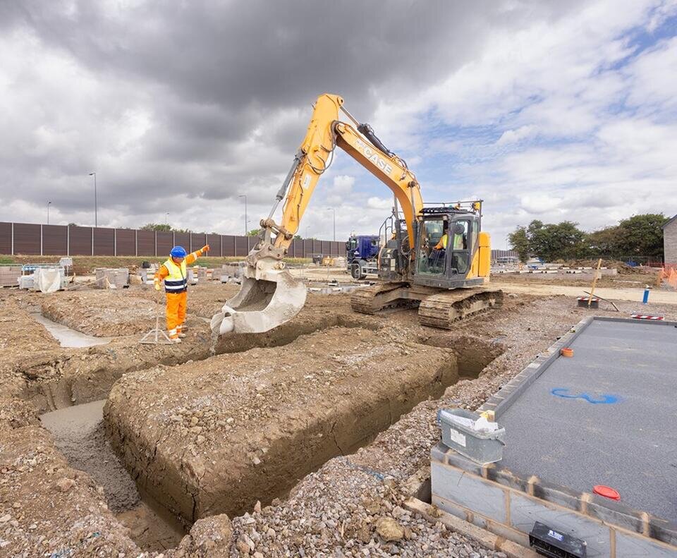 a large digger pouring concrete into a a square in the ground. Next to the digger is a groundsworker in orange high vis clothes and a yellow high vis vest.