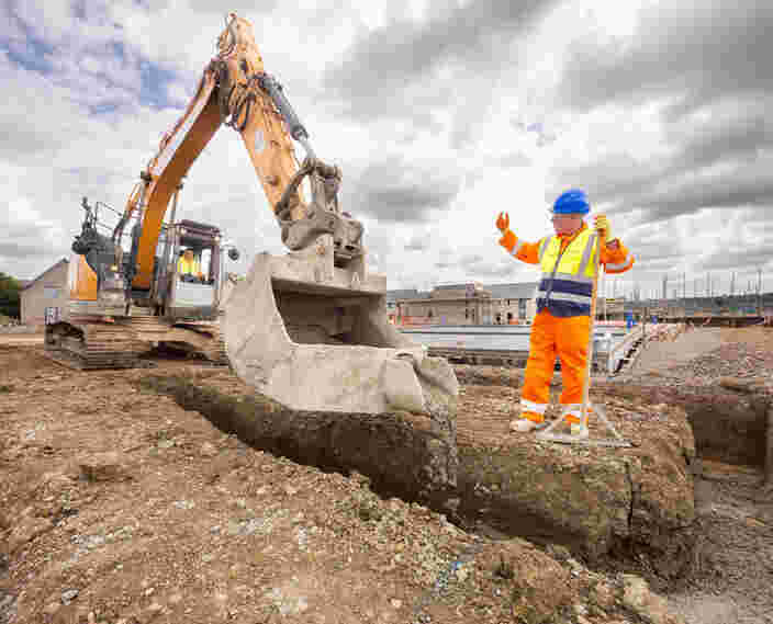 A groundworker is stood on the right directing a large digger which is pouring concrete into the ground