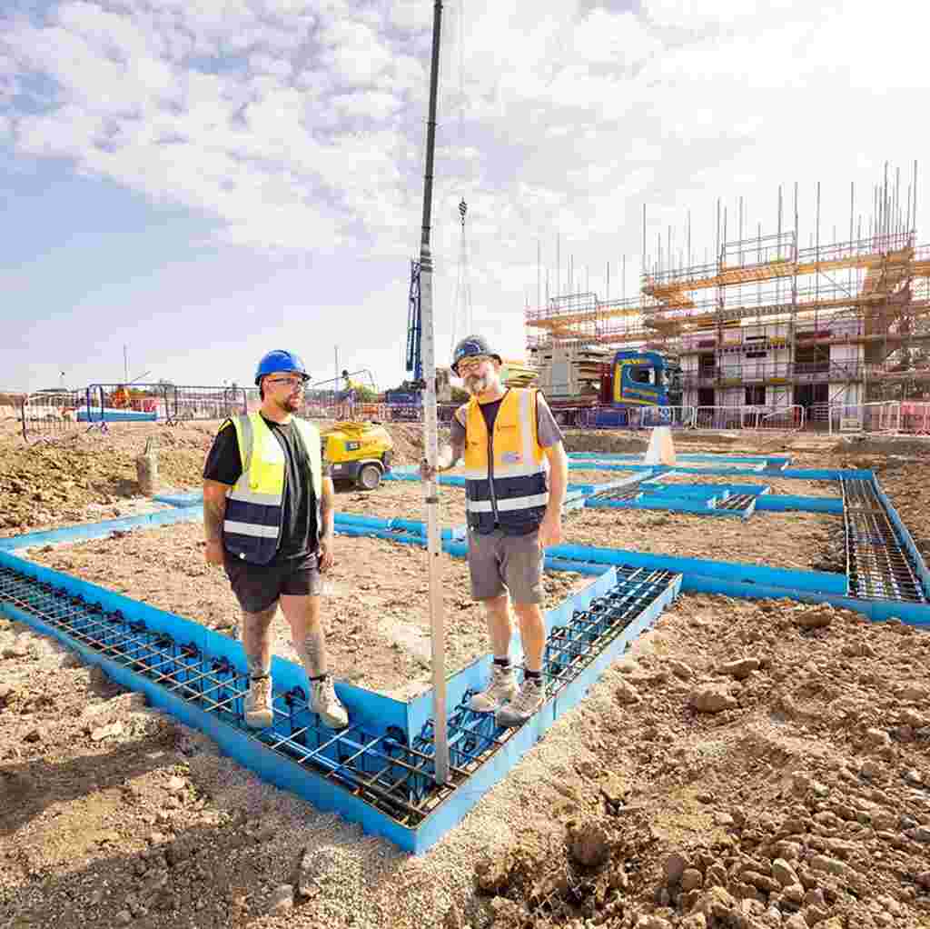 Two groundsworkers in high-visibility vests and hard hats stand on a building site with blue steel foundations. Scaffolding and machinery are visible in the background under a sunny sky.