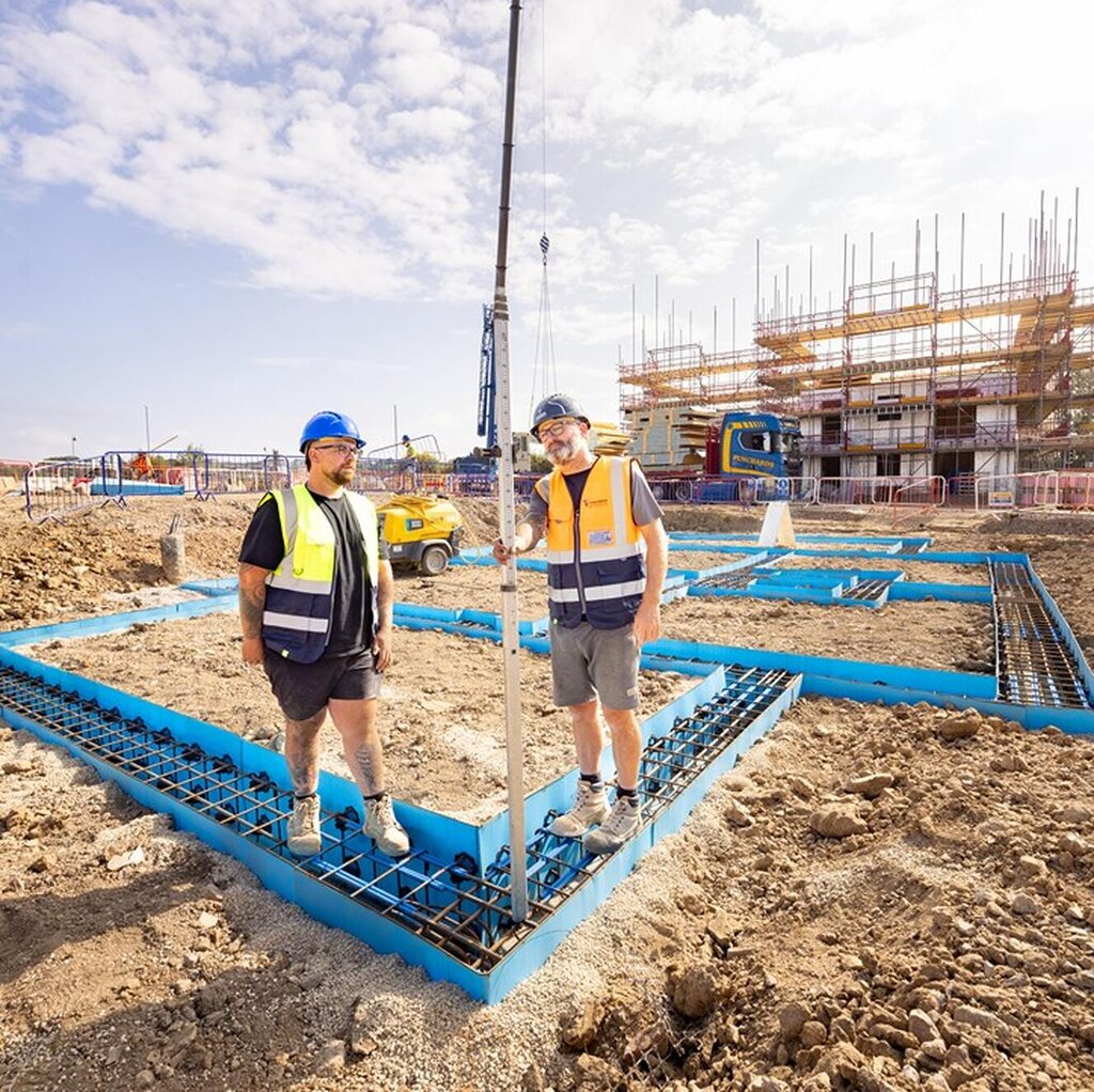 Two groundsworkers in high-visibility vests and hard hats stand on a building site with blue steel foundations. Scaffolding and machinery are visible in the background under a sunny sky.