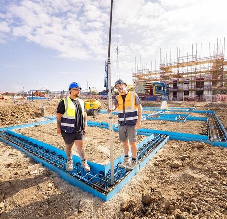 Construction site with two groundsworkers in high vis vests and helmets standing on a square foundation. Behind them are scaffolding and construction vehicles, under a blue sky.