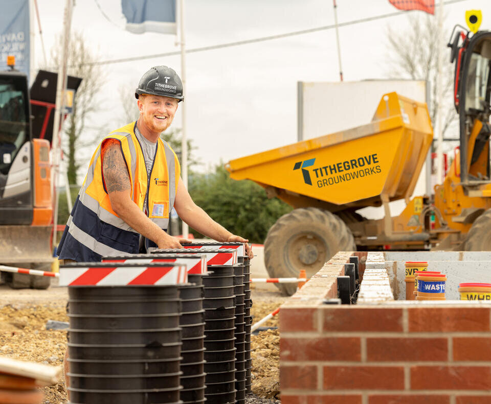 A groundsworker smiles at the camera. He is placing large black cylinders into the ground with red and white tape at the top. In the background is a dumper truck, with tithegrove's logo clearly visible.