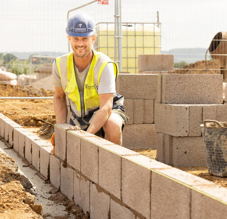 A groundsworker in a high vis vest and blue hard hat lays grey breezeblocks