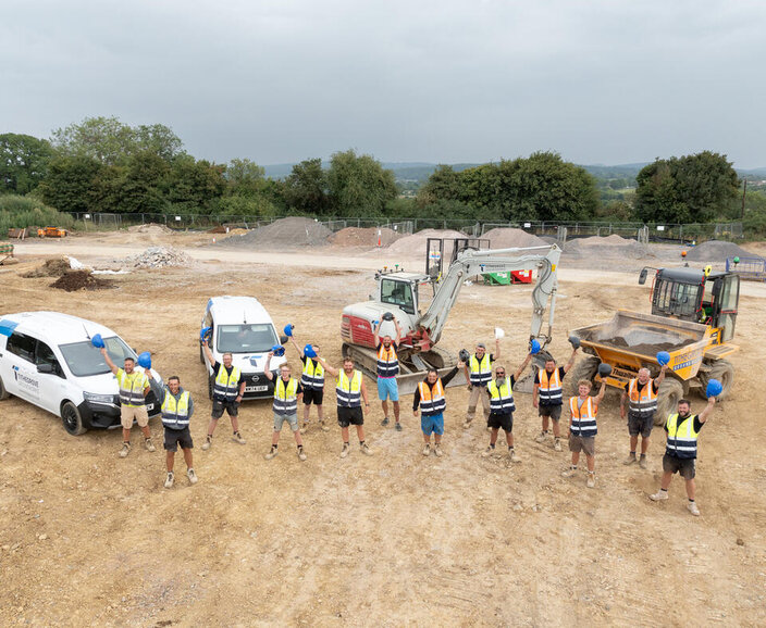 A group of construction workers wearing high-visibility vests holding blue hard hats in the air at a construction site, surrounded by vehicles and machinery.