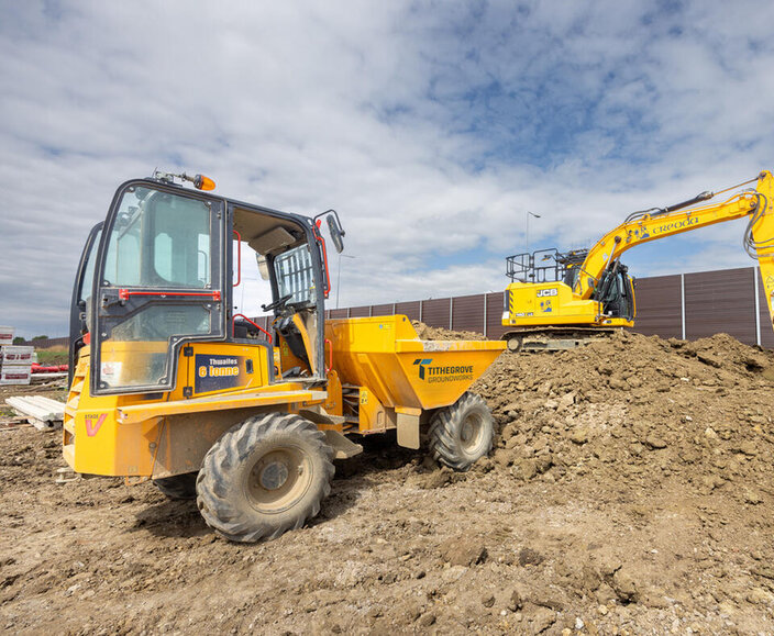 A yellow dumper with a safety shield pours muck onto a pile of earth while a yellow digger spreads the muck