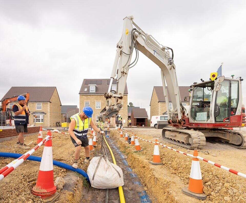 a large digger on the left is manoeuvring a one tonne bag over newly laid utility cables. The cables are exposed. Two groundsworkers are directing the digger.