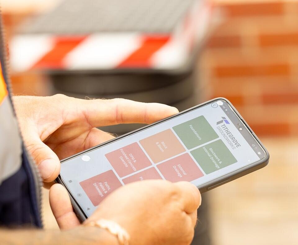A person holds a smartphone displaying a construction management app, with coloured tiles and text. Background shows brickwork and a blurred caution barrier. The setting suggests a worksite, conveying a sense of activity and focus.