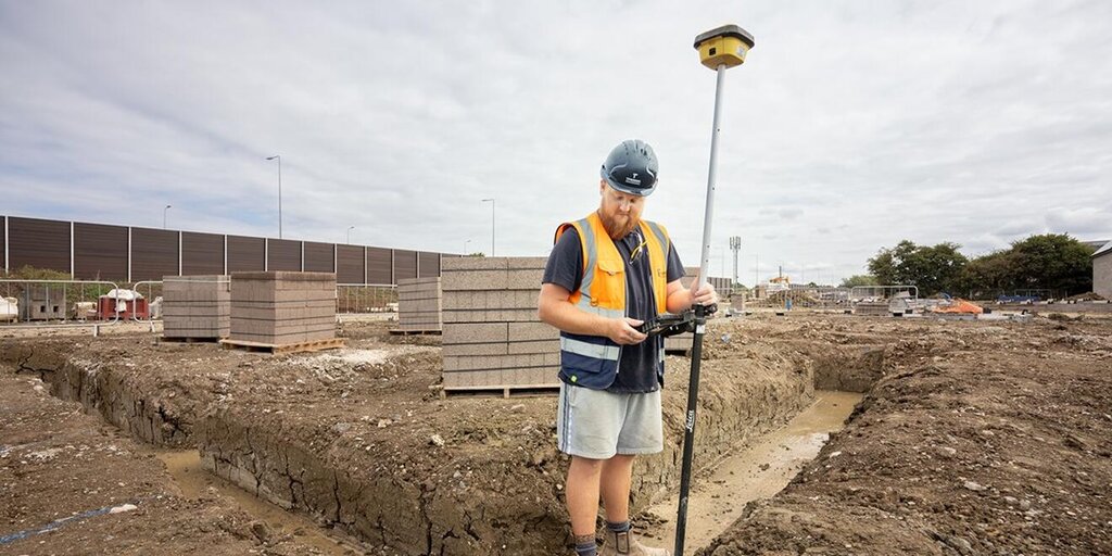 A groundsworker in a hard hat and orange vest holds a device on a muddy site. Piles of bricks are in the background under a cloudy sky.