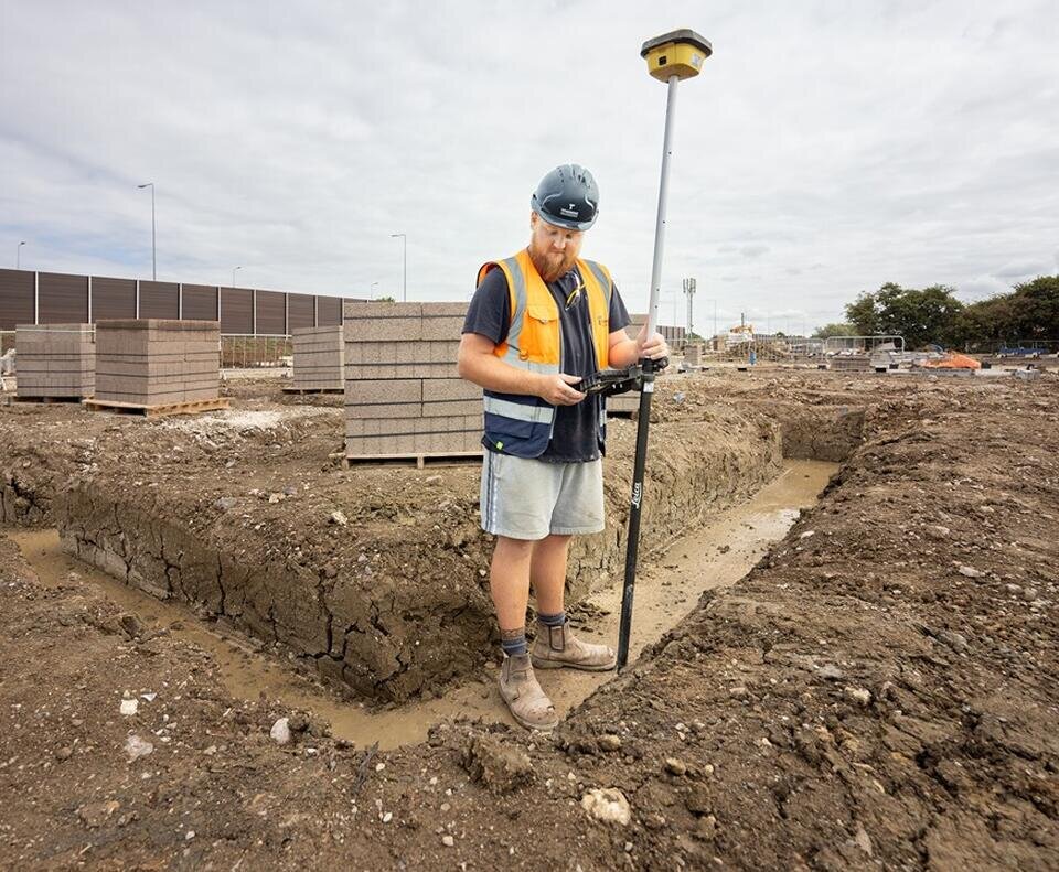 A groundsworker is in the centre of the image. He is stood in a ditch where foundations for a building have been cut out of the earth. He is holding a large metal pole.