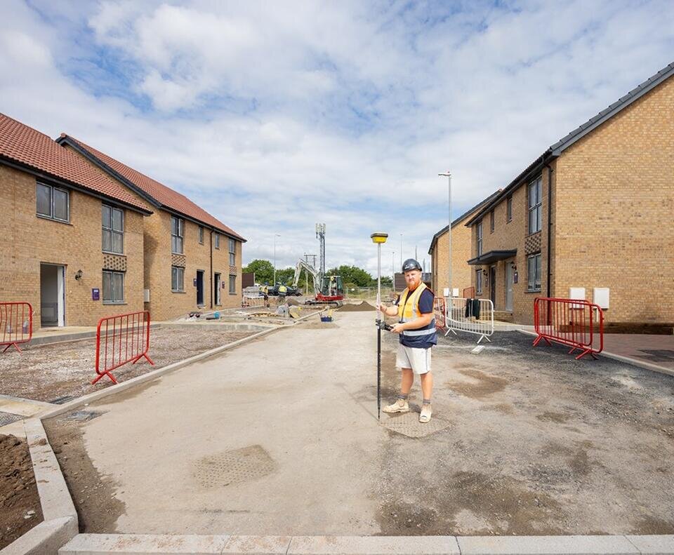 A groundworker stands centre while new build homes are either side. The groundworker is holding a large metal pole.