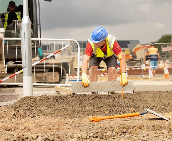 A groundsworker in a blue hat and high vis vest is laying kerbing. Behind him is a man in a digger.
