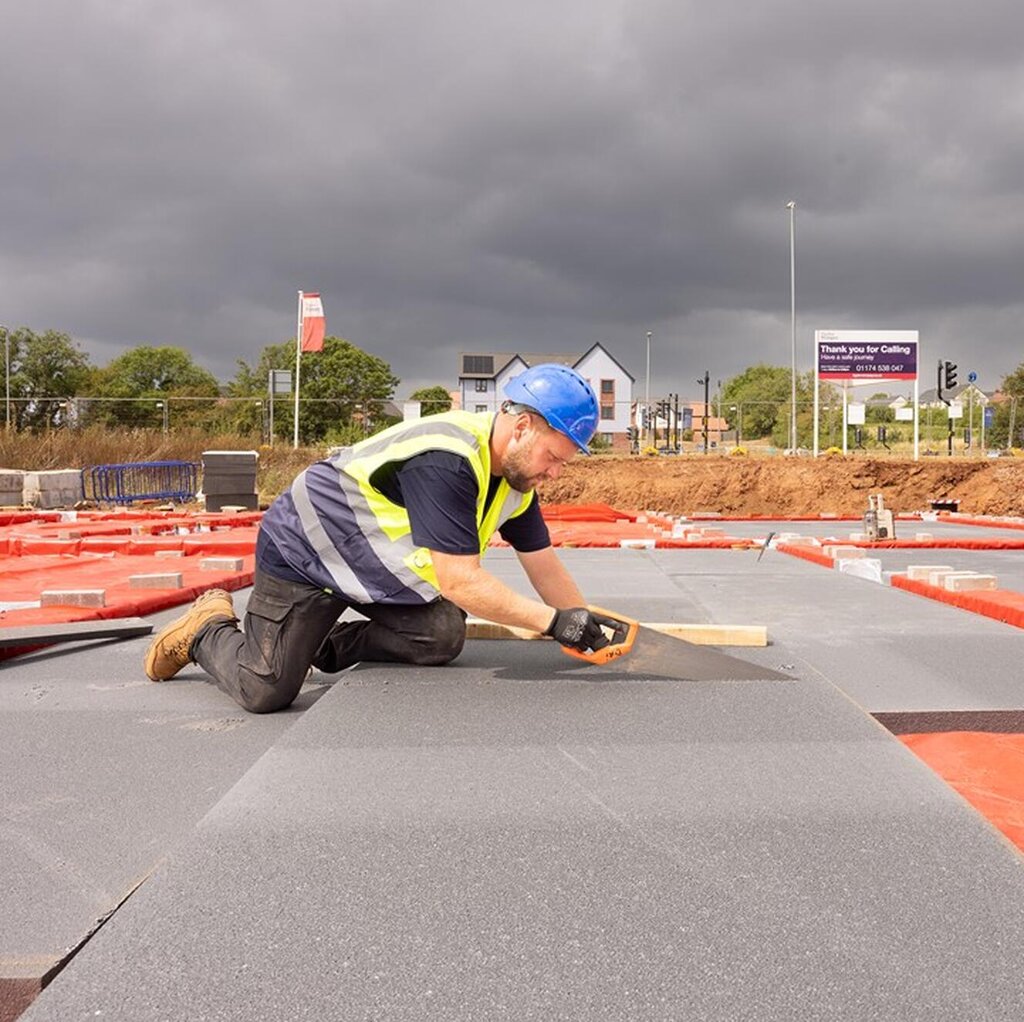 A groundworker is kneeling down with a handsaw. He is cutting boards which will be part of the foundations of a new home.