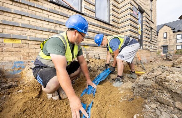 Two groundsworkers are crouched down laying mains services and drainage outside a new build house