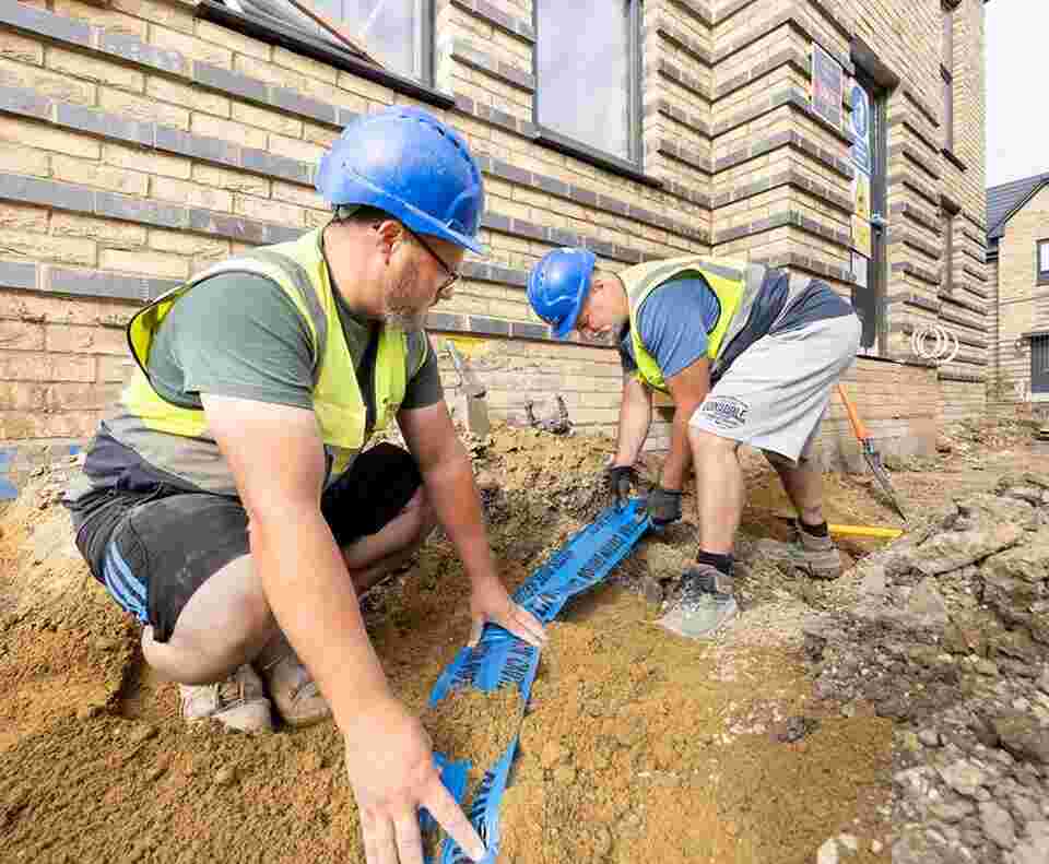 Two groundsworkers are crouched down laying mains services and drainage outside a new build house