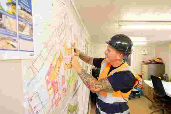 A groundsworker stands and points at a map of a new build site which is pinned on a wall. He is wearing an orange high vis jacket and black hard hat
