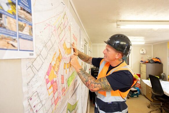 A groundsworker stands and points at a map of a new build site which is pinned on a wall. He is wearing an orange high vis jacket and black hard hat