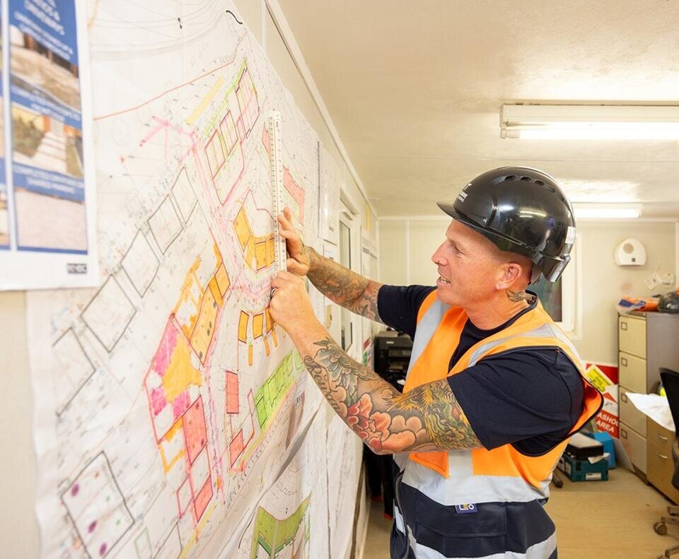 A groundsworker in an orange high vis vest and black hard hat is stood at a large site map which is printed and stuck on a whiteboard.