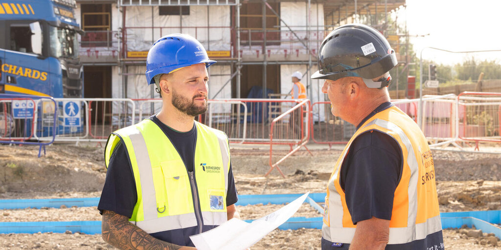 On the left is a groundsworker in an orange high vis, black tshirt and black hard hat. He is holding a metal pole and looking at a large printed document held by a groundworker on the right who is wearing a yellow high vis vest and blue hard hat. The two men are stood on a building site.