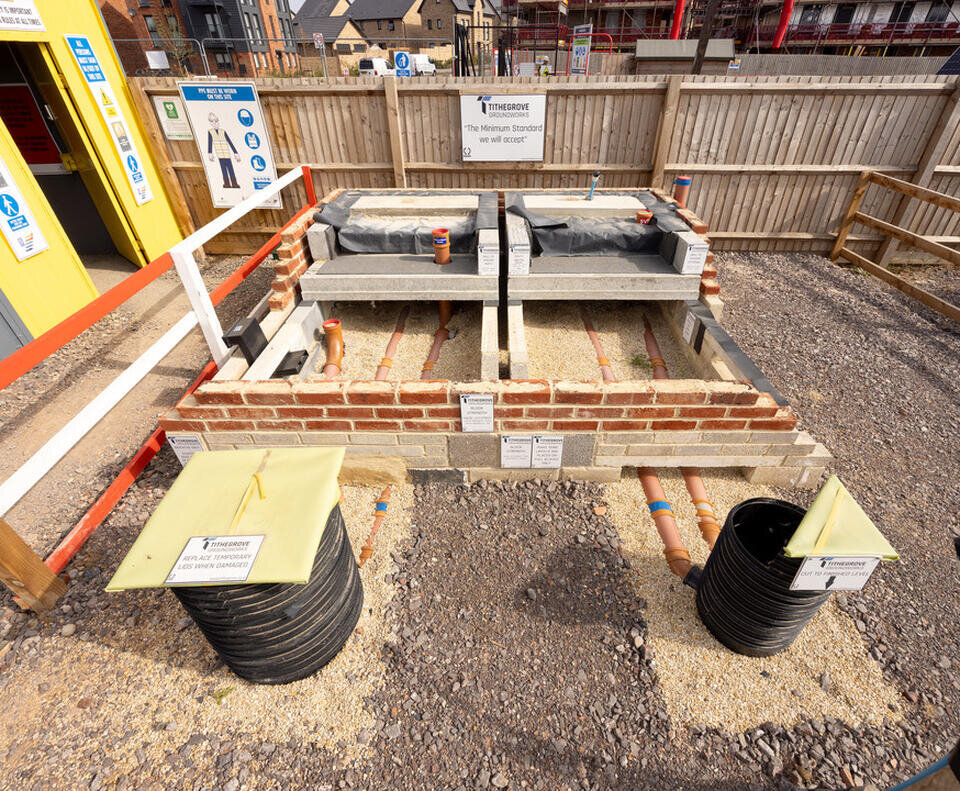 Construction site display of a foundations with exposed pipes and brickwork, surrounded by signage and barriers. A shed and fencing are in the background.
