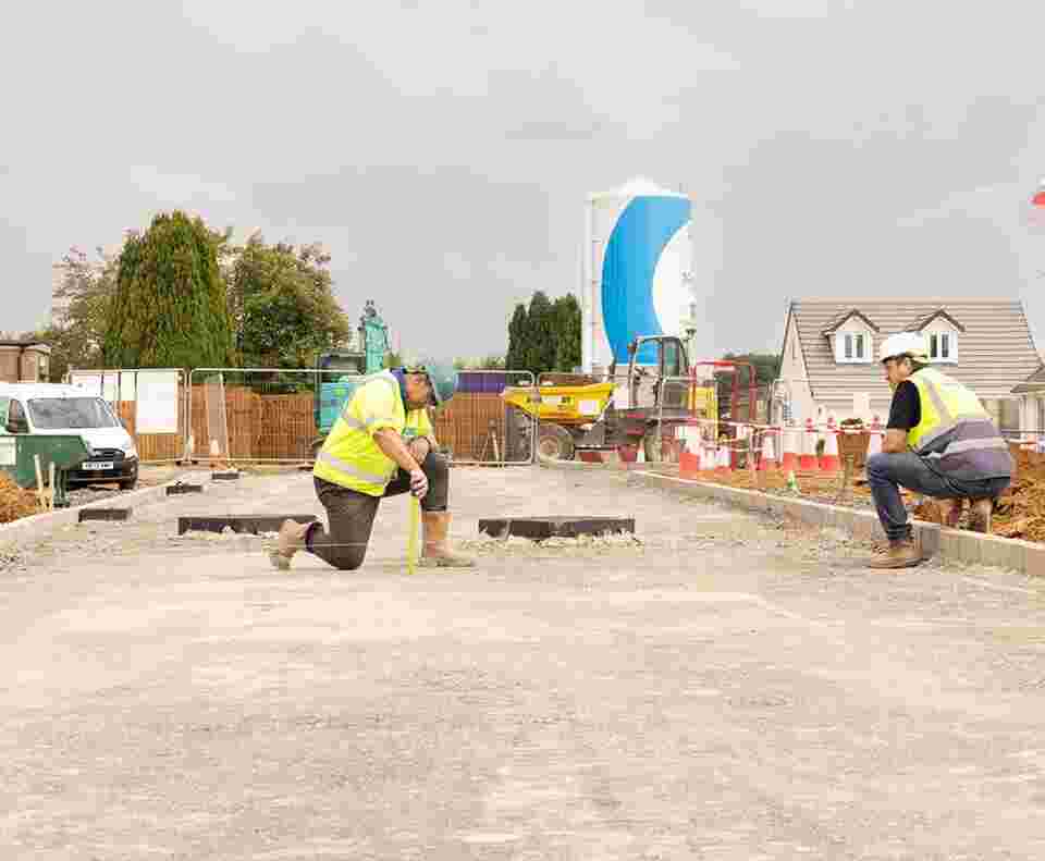 two groundsworkers in high vis vests and hard hats knealing on an unfinished road. They are making sure the kerb is level using string.