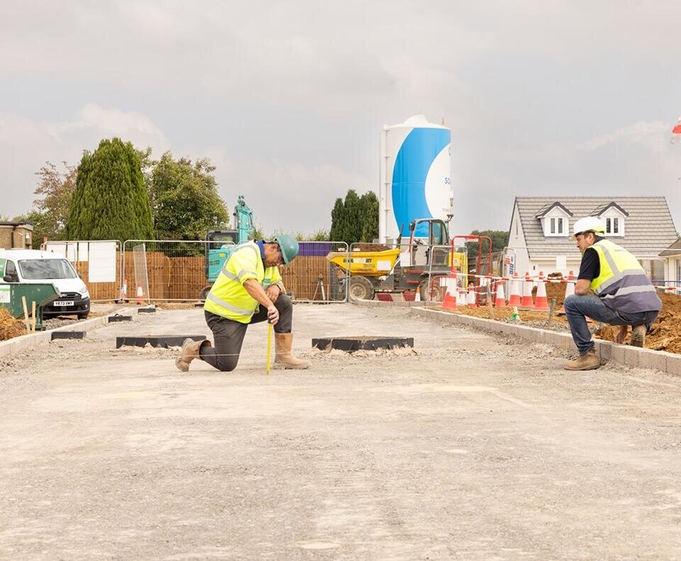 two groundsworkers in high vis vests and hard hats knealing on an unfinished road. They are making sure the kerb is level using string.