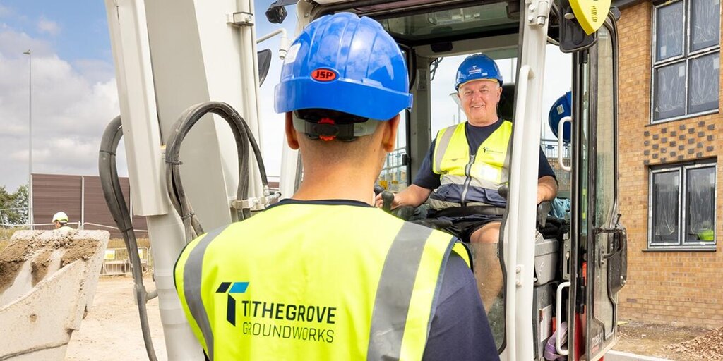 In the background is a groundworker sat in a digger smiling. In the foreground is a groundworker from the back, he is wearing a blue hard hat and yellow high vis vest with tithegrove's logo clearly visibile