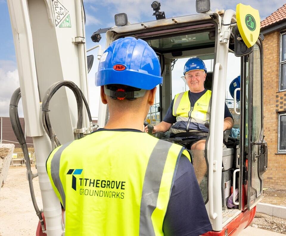 Groundsworkers wearing blue helmets and yellow vests communicate at a site next to a building. One is in an excavator, the other stands outside.